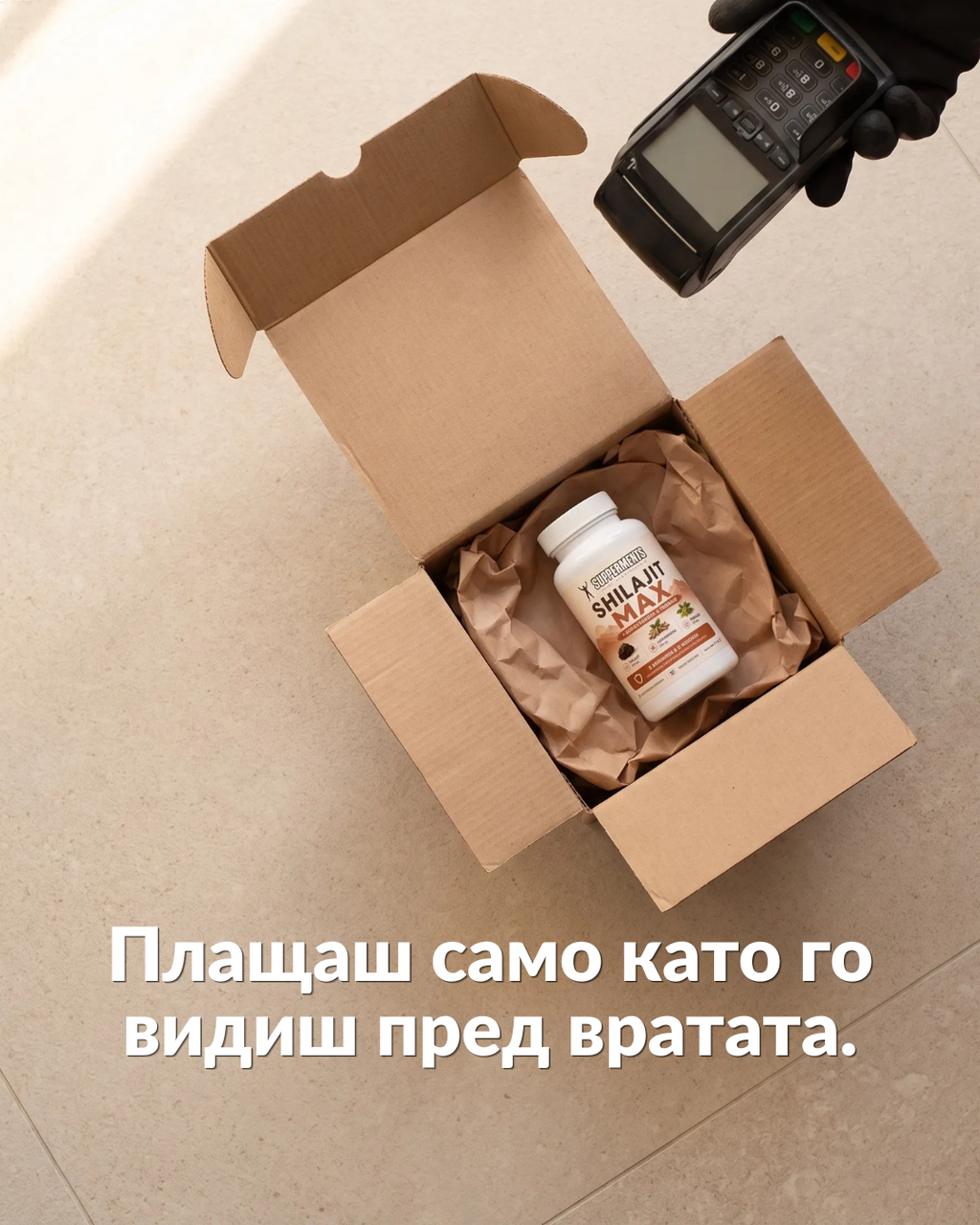An open cardboard box on a light beige floor contains a white supplement bottle wrapped in brown paper. A gloved hand holds a handheld payment terminal above the box.